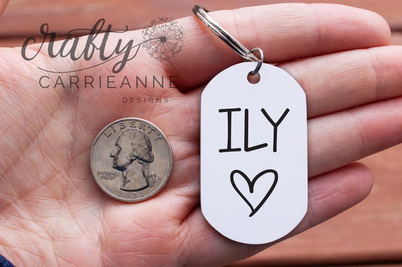 May include: White metal keychain with the text "ILY" and a heart drawn on it. The keychain is being held in a hand.