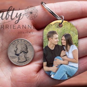 May include: A silver keychain with a photo of a couple in a field. The photo is oval shaped and the keychain is in the palm of a hand.