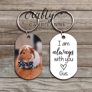 May include: A set of two oval-shaped keychains. One keychain features a photo of a guinea pig wearing a bow tie. The other keychain is white with black text that reads "I am always with you" and a heart with the name "Gus" underneath.
