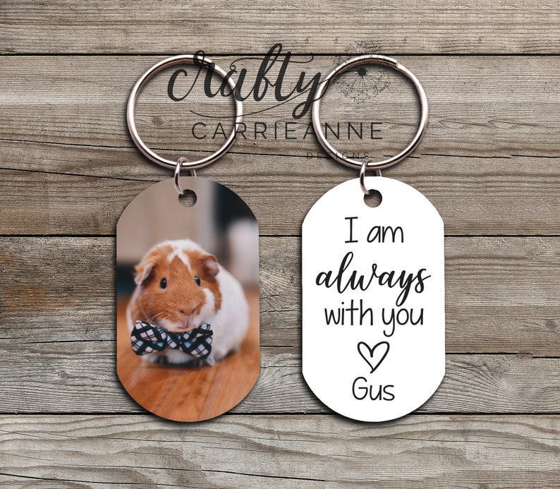 May include: A set of two oval-shaped keychains. One keychain features a photo of a guinea pig wearing a bow tie. The other keychain is white with black text that reads "I am always with you" and a heart with the name "Gus" underneath.