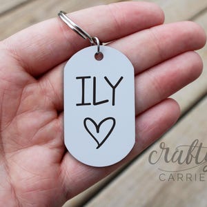 May include: A white metal keychain with a silver key ring. The keychain has the text "ILY" with a heart drawn below it.