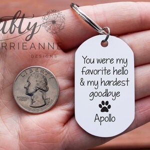 May include: White oval keyring with the text "You were my favourite hello & my hardest goodbye" and a paw print. The name "Apollo" is printed below the paw print. The keyring is held in a hand with a quarter in the background.