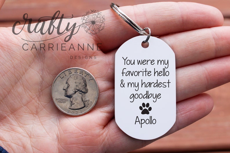 May include: White oval keyring with the text "You were my favourite hello & my hardest goodbye" and a paw print. The name "Apollo" is printed below the paw print. The keyring is held in a hand with a quarter in the background.