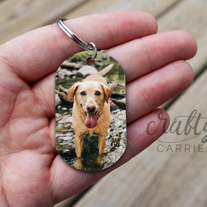 Op de afbeelding: Een gele Labrador Retriever met zijn tong naar buiten stekend staat in een ondiepe beek. De hond zit aan een sleutelhanger met een zilveren ovaalvormig label.