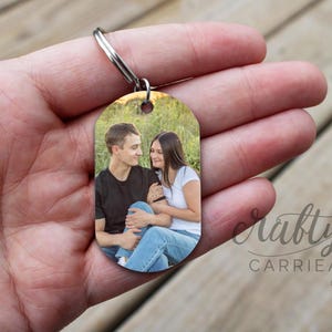 May include: A silver keychain with a photo of a couple in a field. The photo shows a man and a woman smiling at each other. The keychain is oval shaped and has a smooth surface.