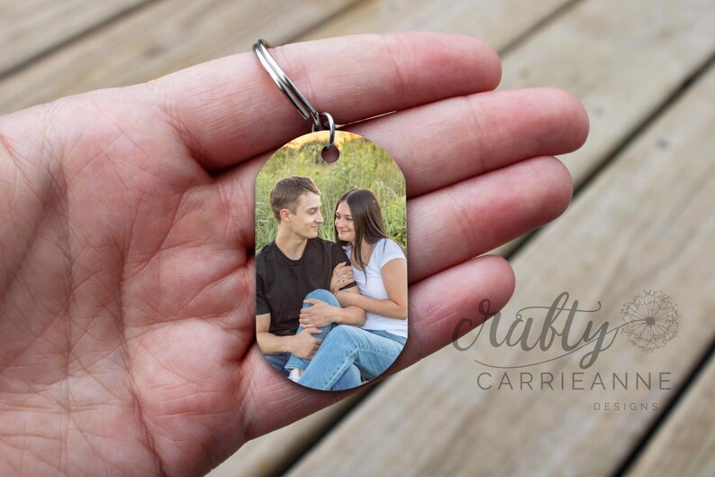 May include: A silver keychain with a photo of a couple in a field. The photo shows a man and a woman smiling at each other. The keychain is oval shaped and has a smooth surface.