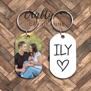 May include: A set of two keychains. One keychain features a photo of a couple in a field. The other keychain is white with black text that says "ILY" with a heart.