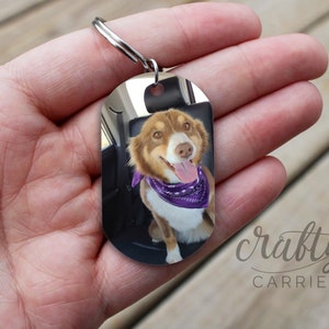 May include: A silver oval-shaped keyring with a photo of a dog wearing a purple bandana. The keyring has a silver ring attached to it. The text "crafty Carrieanne Designs" is visible in the bottom right corner of the image.
