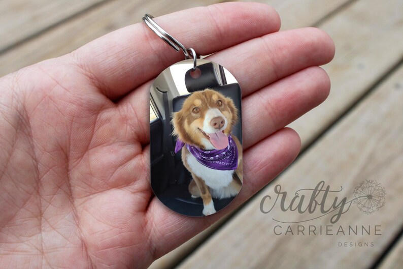 May include: A silver oval-shaped keyring with a photo of a dog wearing a purple bandana. The keyring has a silver ring attached to it. The text "crafty Carrieanne Designs" is visible in the bottom right corner of the image.