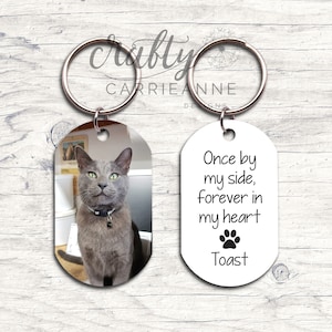 May include: A silver key chain with two oval-shaped tags. One tag has a photo of a gray cat. The other tag has the text "Once by my side, forever in my heart" with a paw print and the name "Toast".