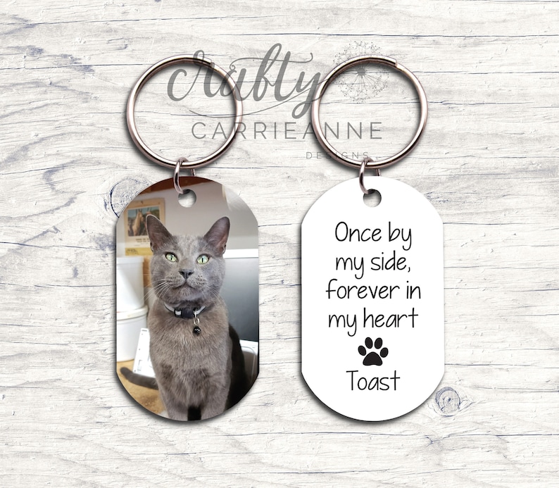 May include: A silver key chain with two oval-shaped tags. One tag has a photo of a gray cat. The other tag has the text "Once by my side, forever in my heart" with a paw print and the name "Toast".