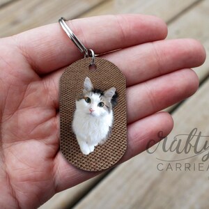 K&ouml;nnte beinhalten: Ein brauner ovaler Schl&uuml;sselanh&auml;nger mit einem Foto einer wei&szlig;en Katze mit braunen und schwarzen Flecken. Die Katze schaut direkt in die Kamera. Der Schl&uuml;sselanh&auml;nger ist aus Holz und hat eine strukturierte Oberfl&auml;che.