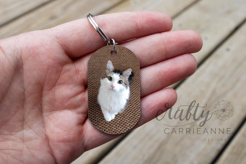 K&ouml;nnte beinhalten: Ein brauner ovaler Schl&uuml;sselanh&auml;nger mit einem Foto einer wei&szlig;en Katze mit braunen und schwarzen Flecken. Die Katze schaut direkt in die Kamera. Der Schl&uuml;sselanh&auml;nger ist aus Holz und hat eine strukturierte Oberfl&auml;che.