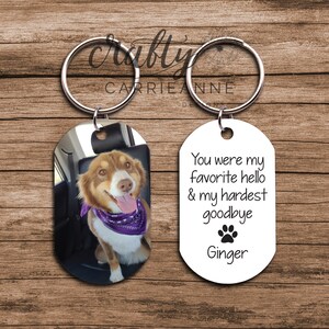 May include: A set of two oval-shaped keychains. One keychain features a photo of a brown and white dog with a purple bandana. The other keychain is white with black text that reads "You were my favourite hello & my hardest goodbye" with a paw print and the name "Ginger".