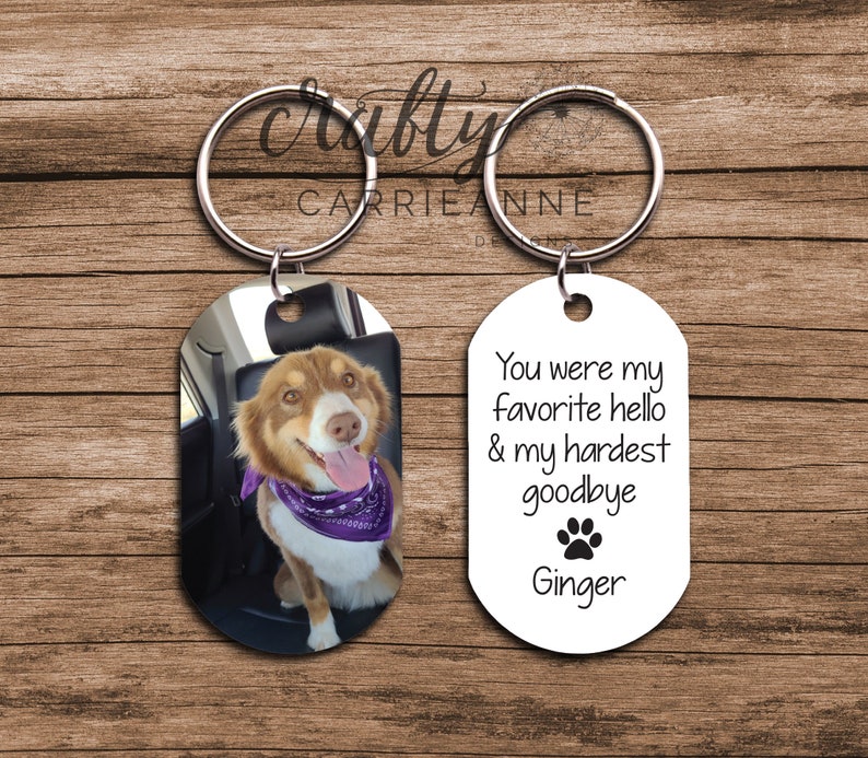 May include: A set of two oval-shaped keychains. One keychain features a photo of a brown and white dog with a purple bandana. The other keychain is white with black text that reads "You were my favourite hello & my hardest goodbye" with a paw print and the name "Ginger".