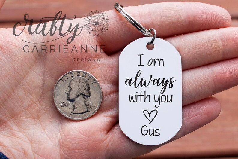 May include: White oval keychain with the text "I am always with you" and a heart symbol. The name "Gus" is written below the heart. The keychain is held in a hand.