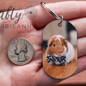 May include: A brown and white guinea pig wearing a black and white bow tie is pictured on a keychain. The keychain is oval shaped and is about the size of a UK 20 pence coin.