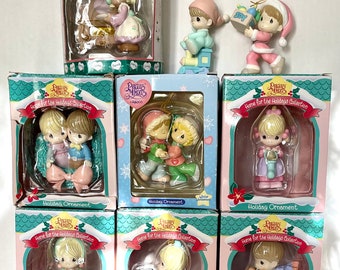 May include: Set of nine Precious Moments Christmas ornaments in various colours and designs. Each ornament features a different scene with the Precious Moments characters. The ornaments are packaged in individual boxes with the words "Home for the Holidays Collection" printed on them.