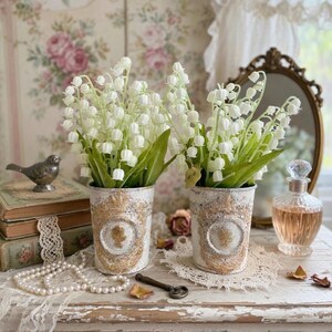 May include: Two white and gold decorative vases filled with lily of the valley flowers. The vases sit on a distressed white table with a vintage mirror and perfume bottle in the background. A string of pearls and antique key are also on the table.