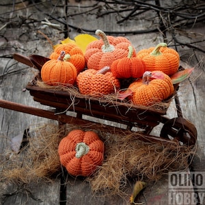 May include: A collection of knitted orange pumpkins in a wooden wheelbarrow. The pumpkins are arranged in a pile with straw and autumn leaves around them.