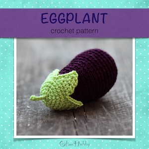 Puede incluir: Juguete amigurumi de berenjena tejido a crochet con un cuerpo morado y un tallo verde. La berenjena está sentada sobre una superficie de madera.