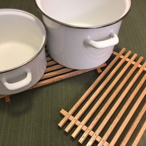 Puede incluir: Dos ollas de esmalte blanco con bordes y asas plateadas que se encuentran sobre una rejilla de madera. La rejilla es de madera marr&oacute;n claro y tiene forma rectangular.