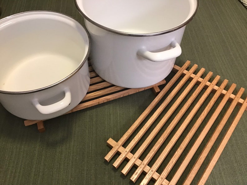 Puede incluir: Dos ollas de esmalte blanco con bordes y asas plateadas que se encuentran sobre una rejilla de madera. La rejilla es de madera marr&oacute;n claro y tiene forma rectangular.
