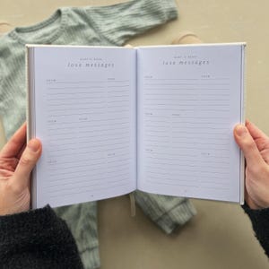 K&ouml;nnte beinhalten: Ein offenes wei&szlig;es Tagebuch mit dem Titel "Baby is here! Love messages" auf dem Vorder- und Hinterdeckel. Das Tagebuch hat linierte Seiten zum Schreiben von Nachrichten.