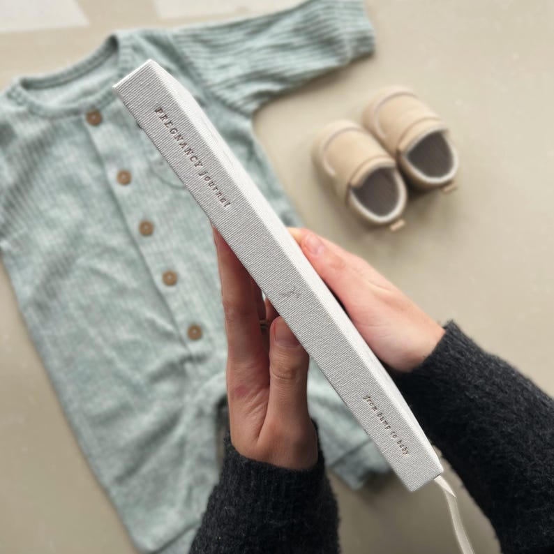 K&ouml;nnte beinhalten: Ein wei&szlig;es Schwangerschaftsjournal mit dem Text "Pregnancy Journey" und "Your heart to hold" auf dem Cover.