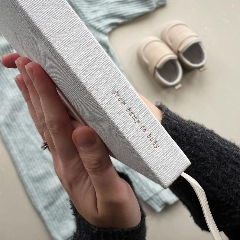 K&ouml;nnte beinhalten: Ein wei&szlig;es, stoffbezogenes Tagebuch mit einem wei&szlig;en Satinband als Lesezeichen. Auf dem Einband steht in Gold "from bump to baby" gedruckt.