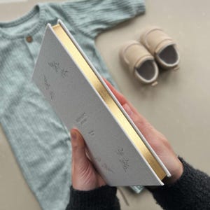 K&ouml;nnte beinhalten: Ein wei&szlig;es Leinen-Tagebuch mit goldenen Kanten und einem goldenen Blumendesign. Das Tagebuch tr&auml;gt den Text "MEMORY JOURNAL" auf dem Einband.