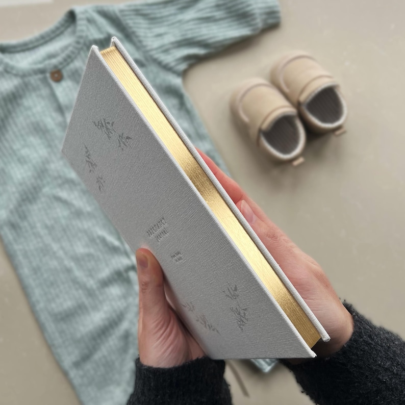 K&ouml;nnte beinhalten: Ein wei&szlig;es Leinen-Tagebuch mit goldenen Kanten und einem goldenen Blumendesign. Das Tagebuch tr&auml;gt den Text "MEMORY JOURNAL" auf dem Einband.