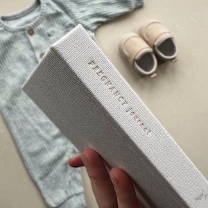 K&ouml;nnte beinhalten: Ein wei&szlig;es, strukturiertes Tagebuch mit dem Aufdruck "Pregnancy Journal" in Gold auf dem Einband.