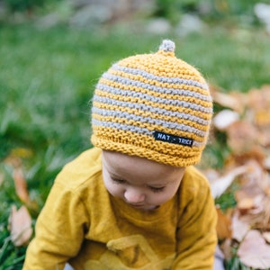May include: A child wearing a yellow jumper and a grey and yellow striped knitted hat with a black label that reads "HAT - TRICK".