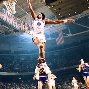 May include: A basketball player wearing a white jersey with blue and red trim, number 6, and the word "Sixers" on the front, jumps to dunk the ball. He is wearing white shorts, blue and white socks, and a knee brace on his right leg. The player is in mid-air, with his right arm extended and his left arm bent at the elbow. The background shows a basketball arena with a crowd of spectators.