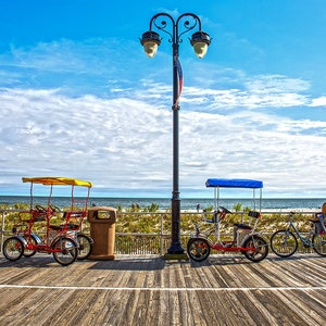 May include: Two red, four-wheeled, pedal-powered surreys with yellow and blue canopies, parked on a wooden boardwalk. A black lamppost with two lights is in the center of the image. The surreys are facing the ocean, which is visible in the background. The sky is blue with white clouds.
