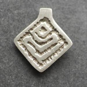 May include: A diamond-shaped silver pendant featuring a raised, textured geometric pattern with a central spiral design. The metal surface has a matte finish with a small bail at the top for attachment, showcasing a distinct, rugged, and hand-crafted appearance.