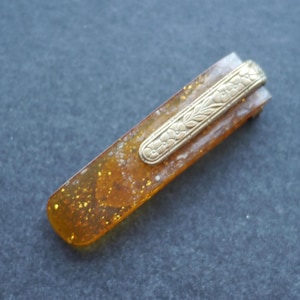 May include: A vintage hair clip with a rectangular amber body speckled with gold glitter. A gold-toned metal floral design decorates the top, adding elegance. This accessory is perfect for adding a vintage flair to any hairstyle.