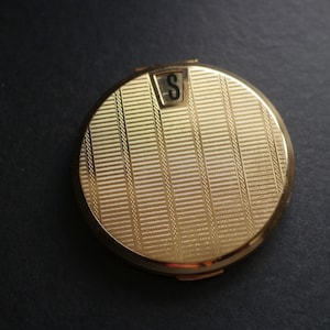 May include: A round gold-toned metal compact mirror featuring a textured surface with vertical stripes and a small rectangular inset at the top containing the black letter S. The compact has a polished finish and a hinged closure mechanism.