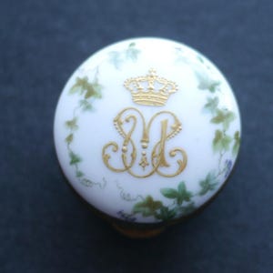 以下が含まれることがあります： 金色の王冠と装飾的なイニシャルが付いた、白い丸い宝石箱の蓋。緑と紫の花柄が金色のデザインを囲んでいます。蓋は金色のベースにセットされています。