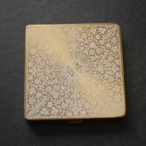Könnte beinhalten: Ein quadratisches Puderetui aus goldfarbenem Metall mit einem filigranen, geätzten weißen Blumenmuster auf der Oberseite. Das Design zeigt eine dichte, symmetrische Anordnung von Blättern und Blüten auf einer goldfarbenen Metalloberfläche.