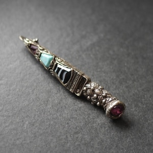 May include: A silver-toned metal pendant shaped like a tapered wand or dagger. The handle features a braided texture, while the blade section is inlaid with a purple stone, a light blue stone, and a black and white striped stone, ending in a purple gem.
