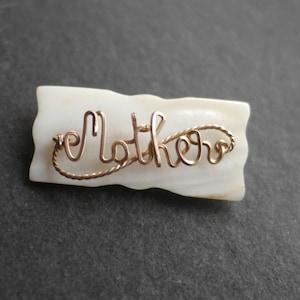 May include: A gold-colored wire brooch spelling "Mother" in cursive script, affixed to a rectangular, off-white, textured base. The brooch is set against a dark gray background.