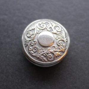 May include: A circular, silver-coloured container with an elaborate floral pattern. The lid has a central disc and detailed, swirling designs. The container is likely metal and has a polished finish.