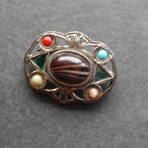 May include: An oval-shaped metal brooch featuring an openwork frame with a central dark brown and white striped cabochon. The piece is accented with a red bead, a turquoise bead, two green triangular inlays, and two small off-white round beads.