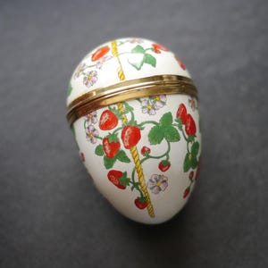 Puede incluir: Un joyero con forma de huevo con una base blanca que presenta un patrón de fresas rojas, hojas verdes, flores rosas y un diseño de cuerda amarilla. La caja tiene un borde de metal dorado que separa las mitades superior e inferior.