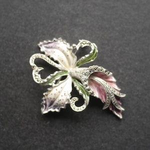 May include: A floral brooch with a silver-tone metal base, featuring white, pink, and green enamel detailing. The brooch is shaped like a stylized flower with marcasite accents, creating a vintage aesthetic.