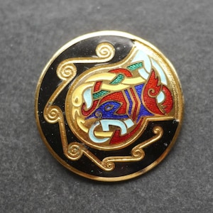 Puede incluir: Un botón redondo de tono dorado con un fondo de esmalte negro y un motivo circular central. El diseño incluye líneas doradas intrincadas y onduladas con un relleno de esmalte vibrante en tonos rojo, azul, verde y azul claro, creando un patrón complejo.