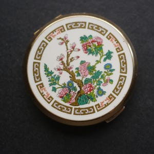 May include: A round gold-tone metal compact mirror featuring a white enamel top with a decorative gold geometric border. The centre displays a colourful illustration of a flowering tree with pink and blue blossoms and green leaves on brown branches.