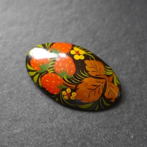 May include: An oval-shaped brooch with a black background featuring a hand-painted design of three bright red strawberries with green stems, brown leaves, and small yellow flowers with red centres. The surface has a glossy, smooth finish.